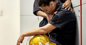 Side view of late 30s woman in uniform sitting on bench in fire station locker room with helmet in lap, head down, and eyes closed.